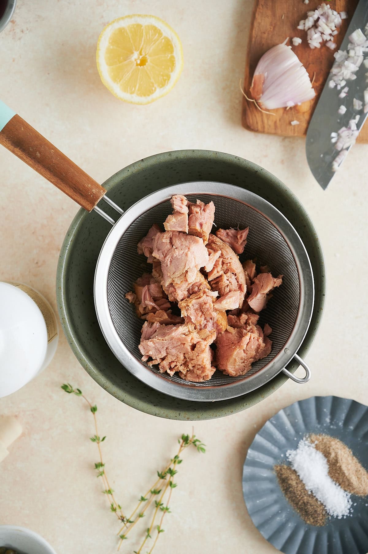 Chunks of tuna sit in a mesh strainer over a bowl—perfect for making Italian tuna salad. Nearby, a halved lemon, chopped shallots, garlic, fresh herbs, and a plate with salt and pepper are neatly arranged on a light surface.
