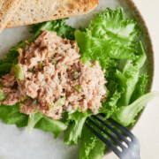 A plate with a serving of tuna salad on a bed of leafy green lettuce, two slices of toasted bread, and a black fork resting on the plate.