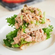 Two triangles of toasted white bread topped with leafy green lettuce and a generous serving of tuna salad are stacked on a white plate. A sliced tomato is visible in the blurred background.