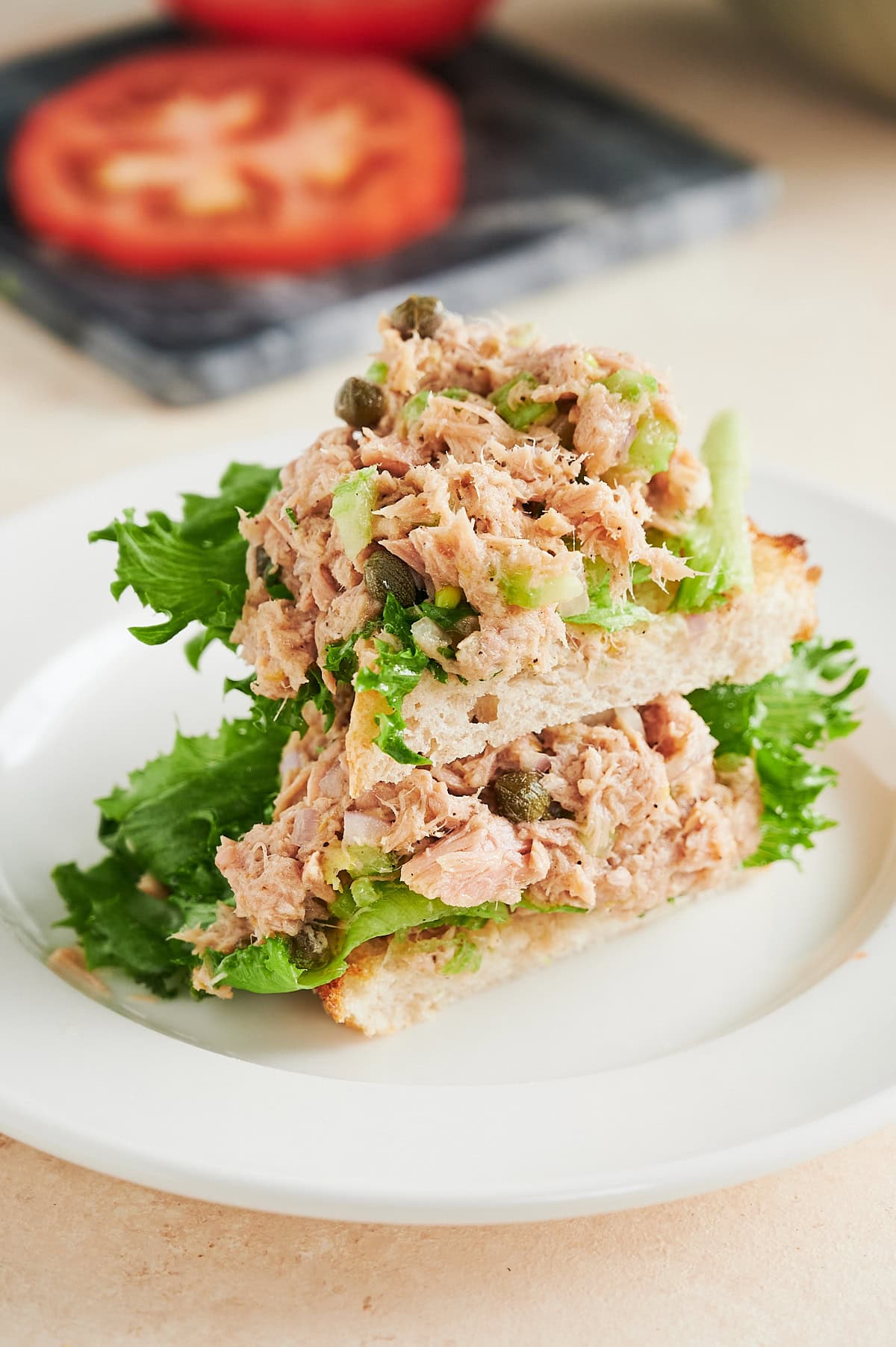 Two triangles of toasted white bread topped with leafy green lettuce and a generous serving of tuna salad are stacked on a white plate. A sliced tomato is visible in the blurred background.