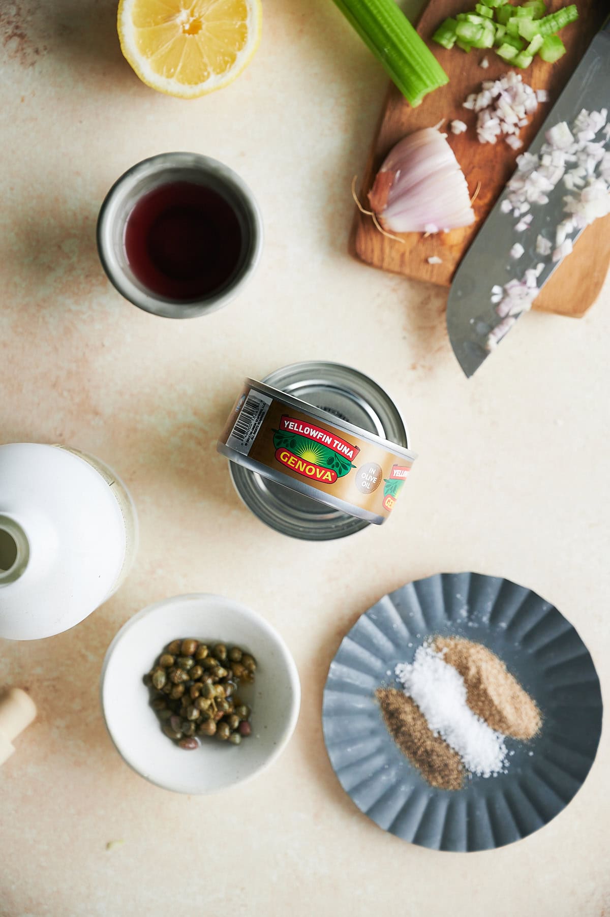 A can of Genova yellowfin tuna on a counter surrounded by a halved lemon, chopped celery, a garlic clove, minced shallots, a glass of red liquid, a bottle, a bowl of capers, and a plate with spices and salt.