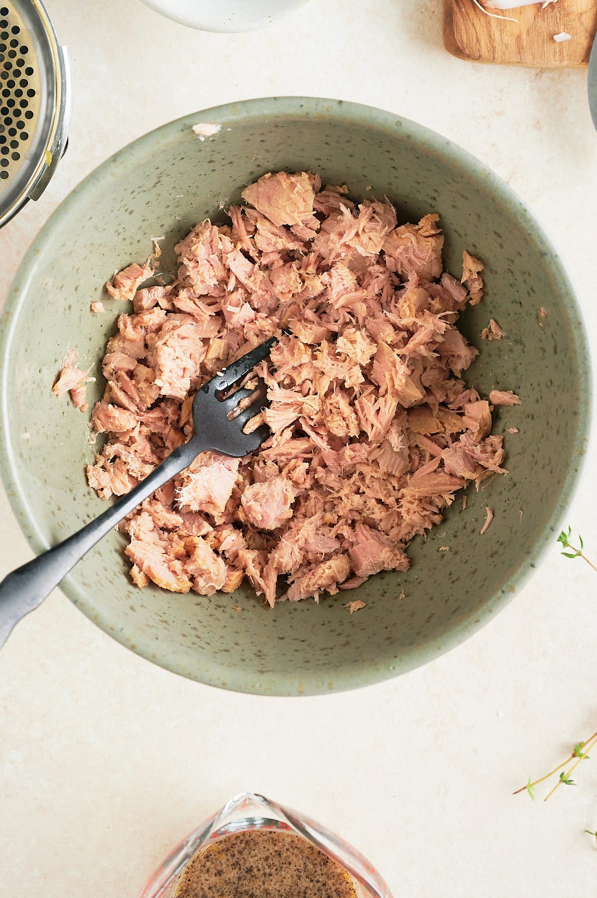 A green bowl filled with flaked tuna and a black fork resting inside. The bowl is on a light-colored surface with part of a spice grinder and a glass measuring cup visible nearby.