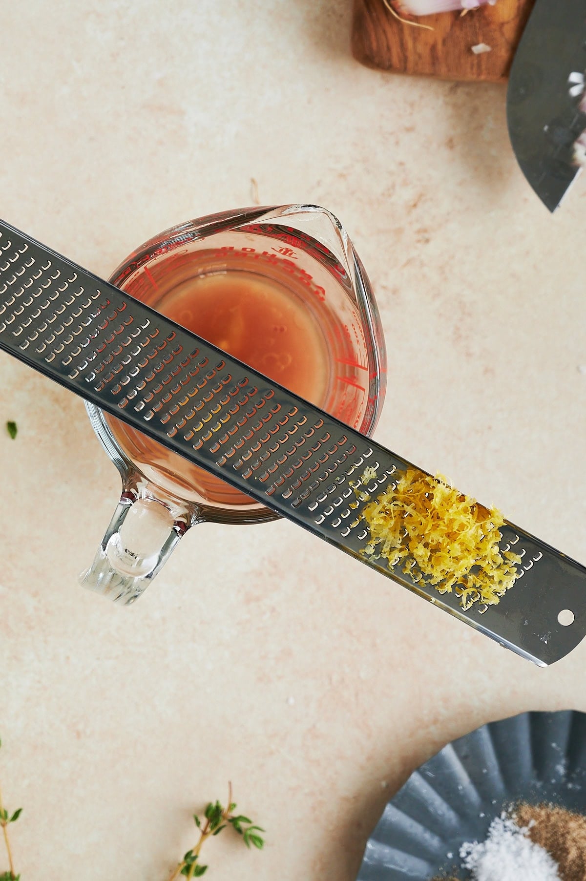 A metal zester with grated lemon zest rests over a glass measuring cup containing a light brown liquid, placed on a beige countertop.