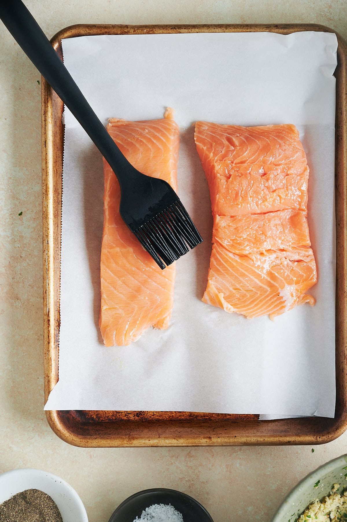 Two raw salmon fillets on a parchment-lined baking sheet being brushed with a black silicone brush.