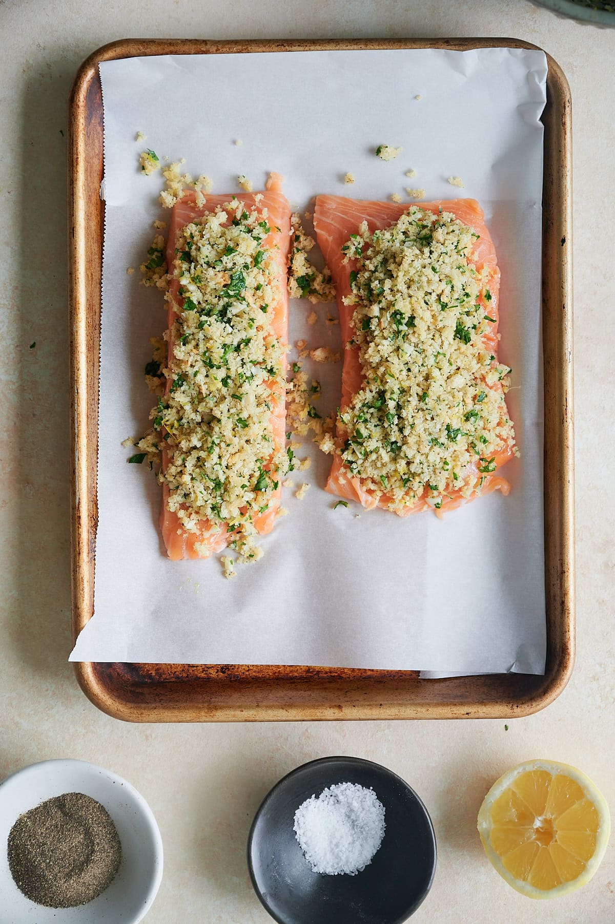 Two raw salmon fillets on a parchment-lined baking tray, topped with a breadcrumb and herb mixture. Ingredients including pepper, salt, and half a lemon are placed nearby.