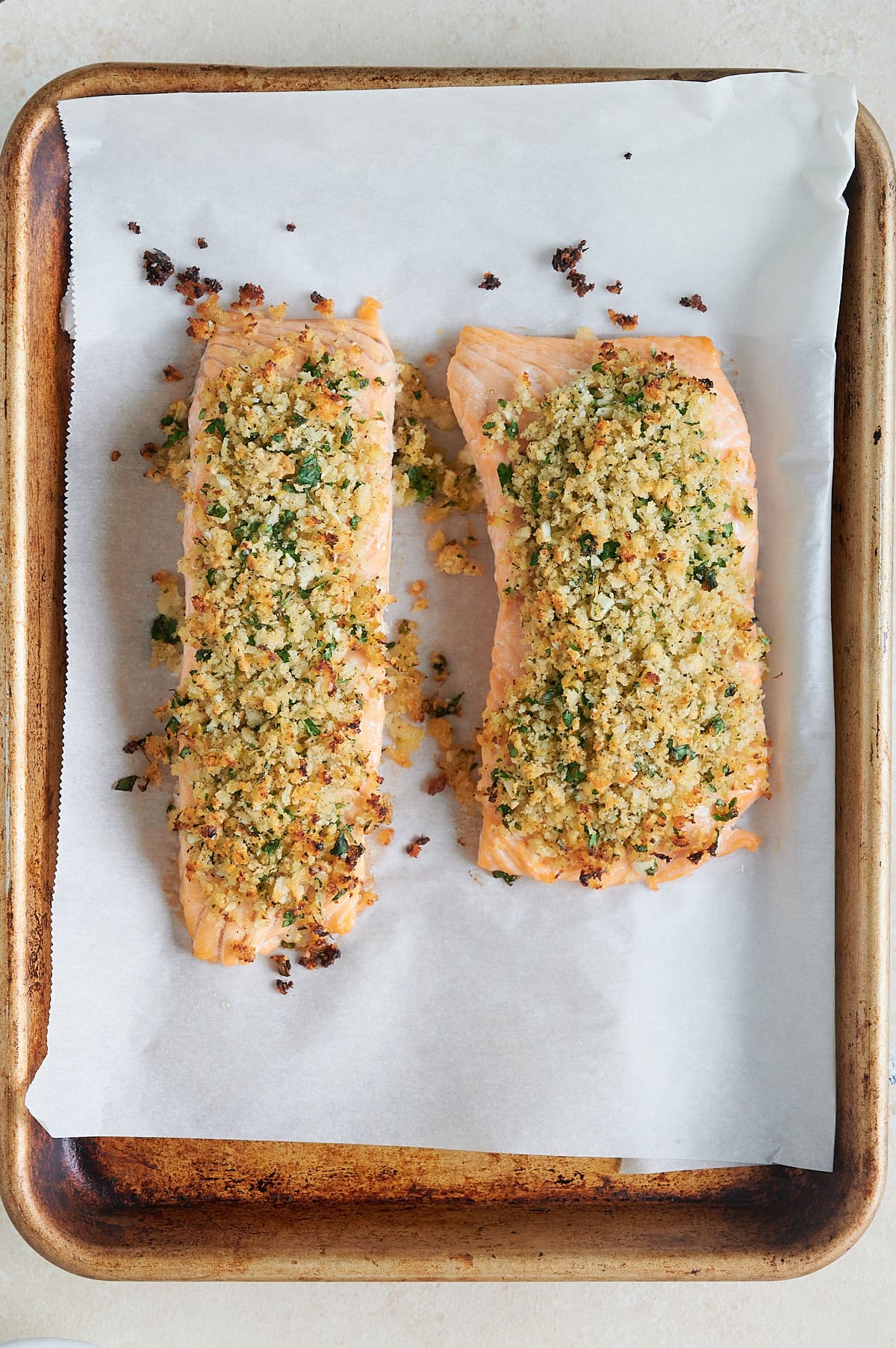 Overhead view of two baked salmon fillets topped with a golden breadcrumb and herb crust on a parchment-lined baking sheet.