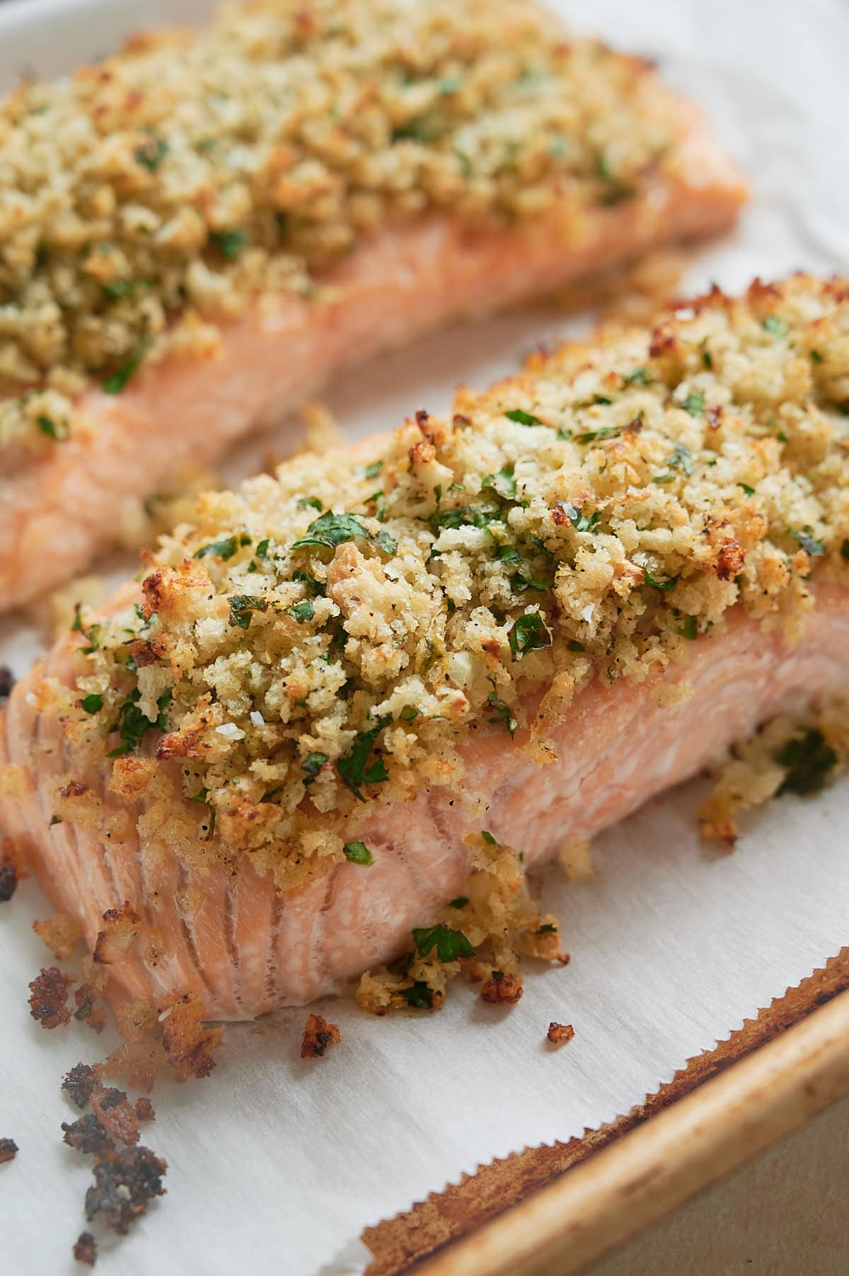 Two baked salmon fillets topped with a golden, herb and breadcrumb crust sit on an angle on a parchment-lined baking sheet.