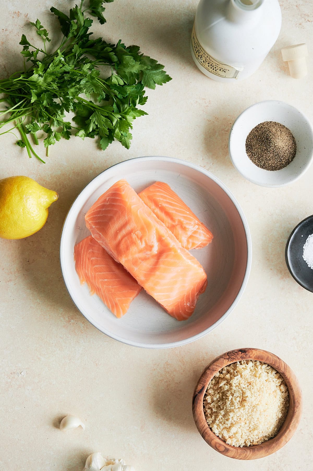 Raw salmon fillets in a bowl surrounded by parsley, a lemon, ground pepper, salt, panko breadcrumbs, garlic cloves, and a bottle of oil on a light countertop.