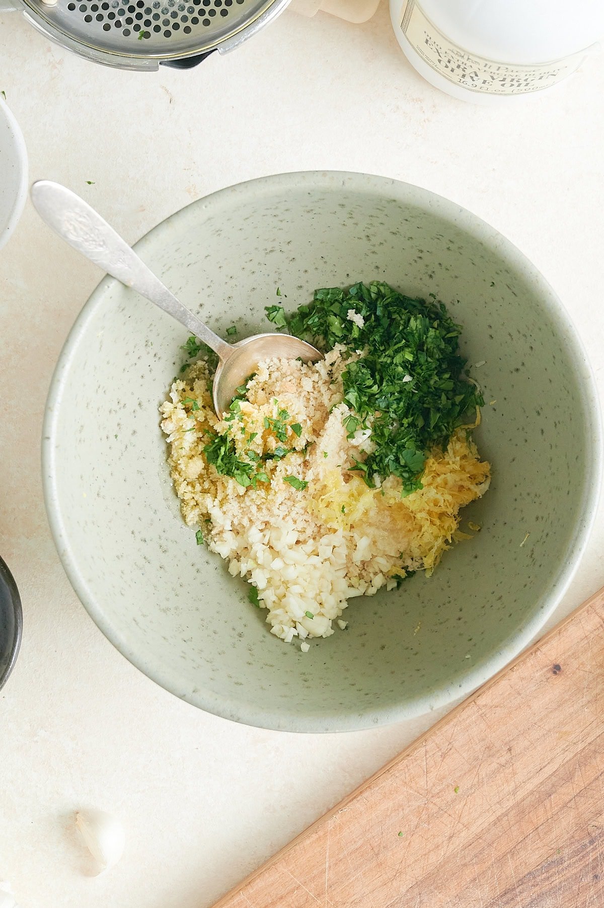 A bowl contains chopped parsley, minced garlic, breadcrumbs, grated cheese, and lemon zest with a spoon inside.