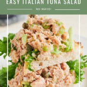 Two open-faced sandwiches on white bread are topped with Italian tuna salad, leafy greens, celery, and capers. Text overlay reads, “Easy Italian Tuna Salad, No Mayo!” Sliced tomatoes are blurred in the background.