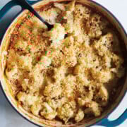 A round blue baking dish filled with baked cauliflower topped with a golden brown breadcrumb crust sits on a light surface. A black serving spoon rests in the dish.