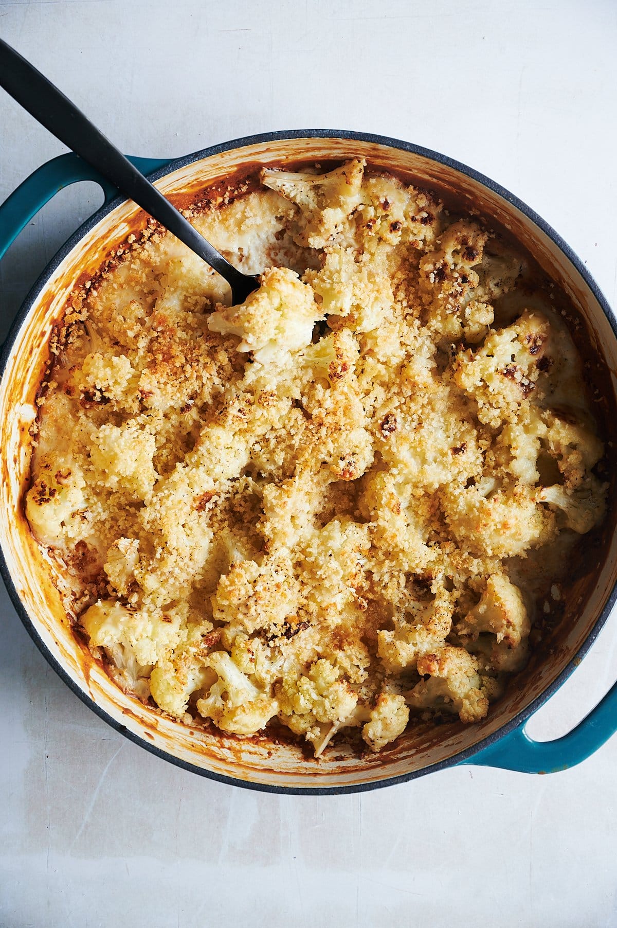 A round blue baking dish filled with baked cauliflower topped with a golden brown breadcrumb crust sits on a light surface. A black serving spoon rests in the dish.