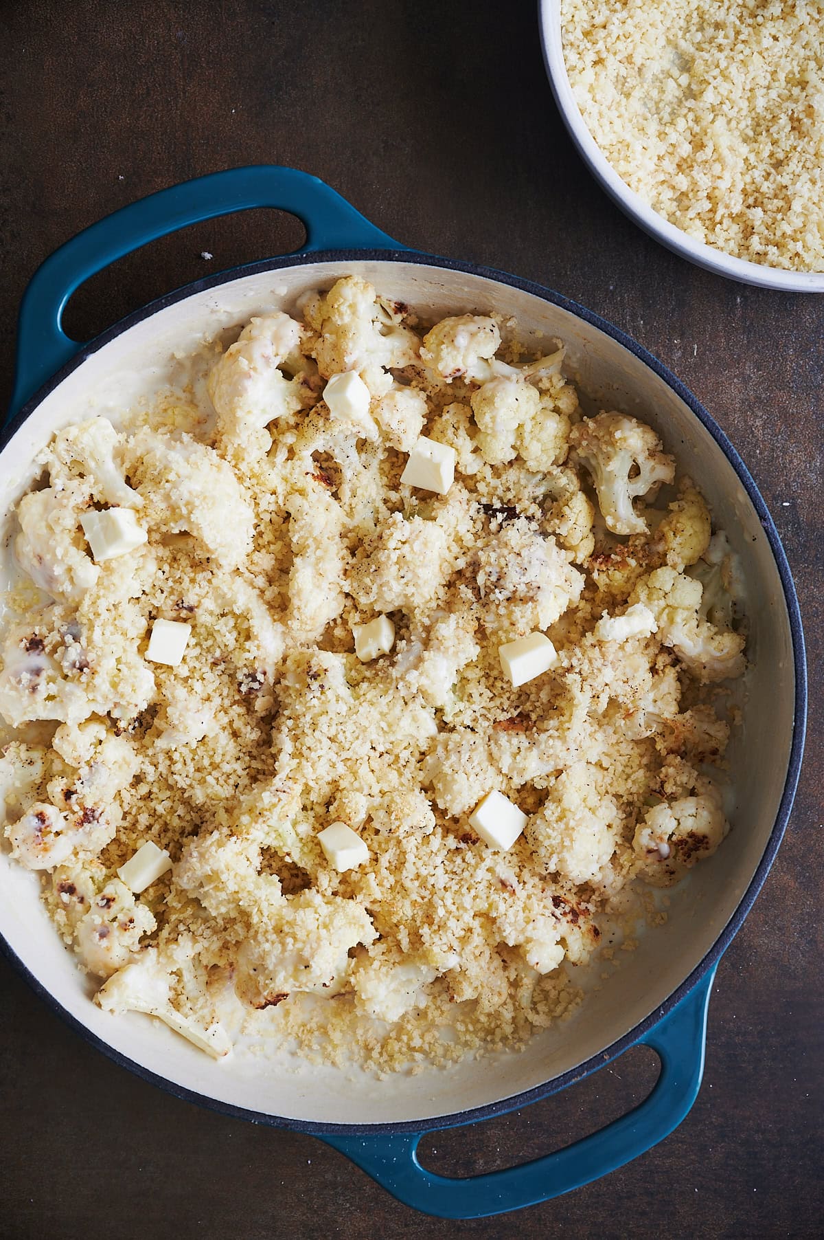 A round baking dish filled with cauliflower florets covered in breadcrumbs, with small cubes of butter scattered on top with a bowl of breadcrumbs visible in the background on a dark surface.