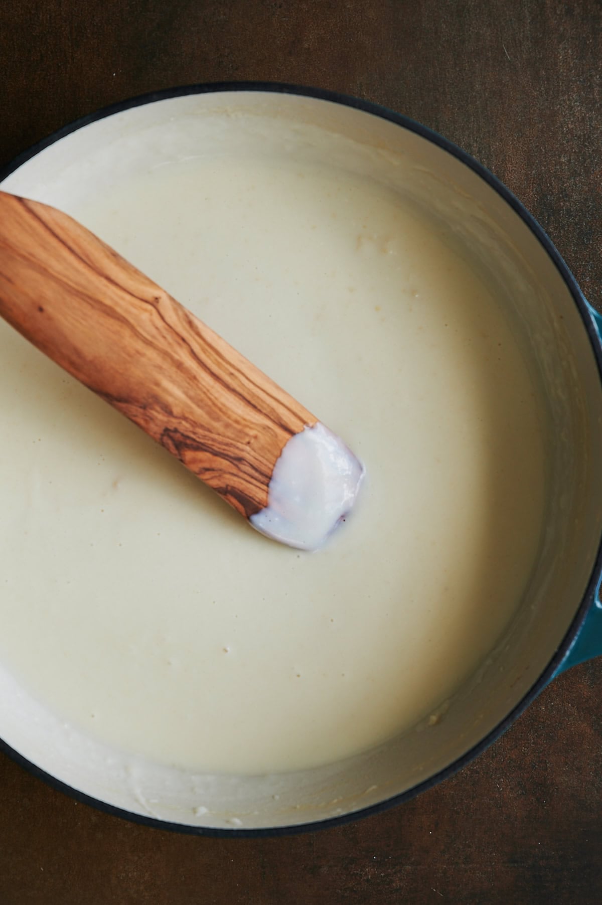 A wooden spoon resting in a pot of creamy white sauce, which has a smooth texture and thick consistency.