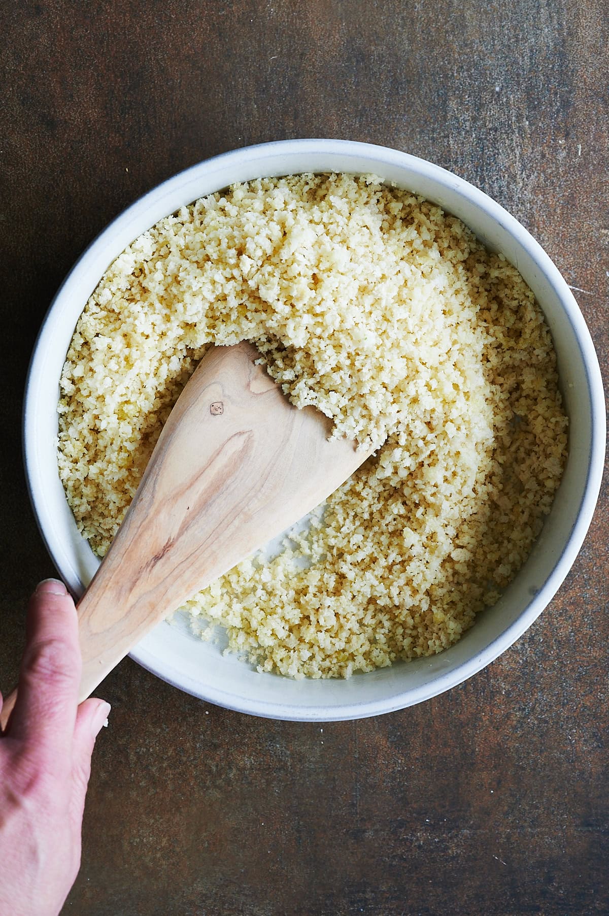 A hand holding a wooden spatula stirs a white bowl filled with cooked couscous or a similar grain, placed on a dark brown surface.