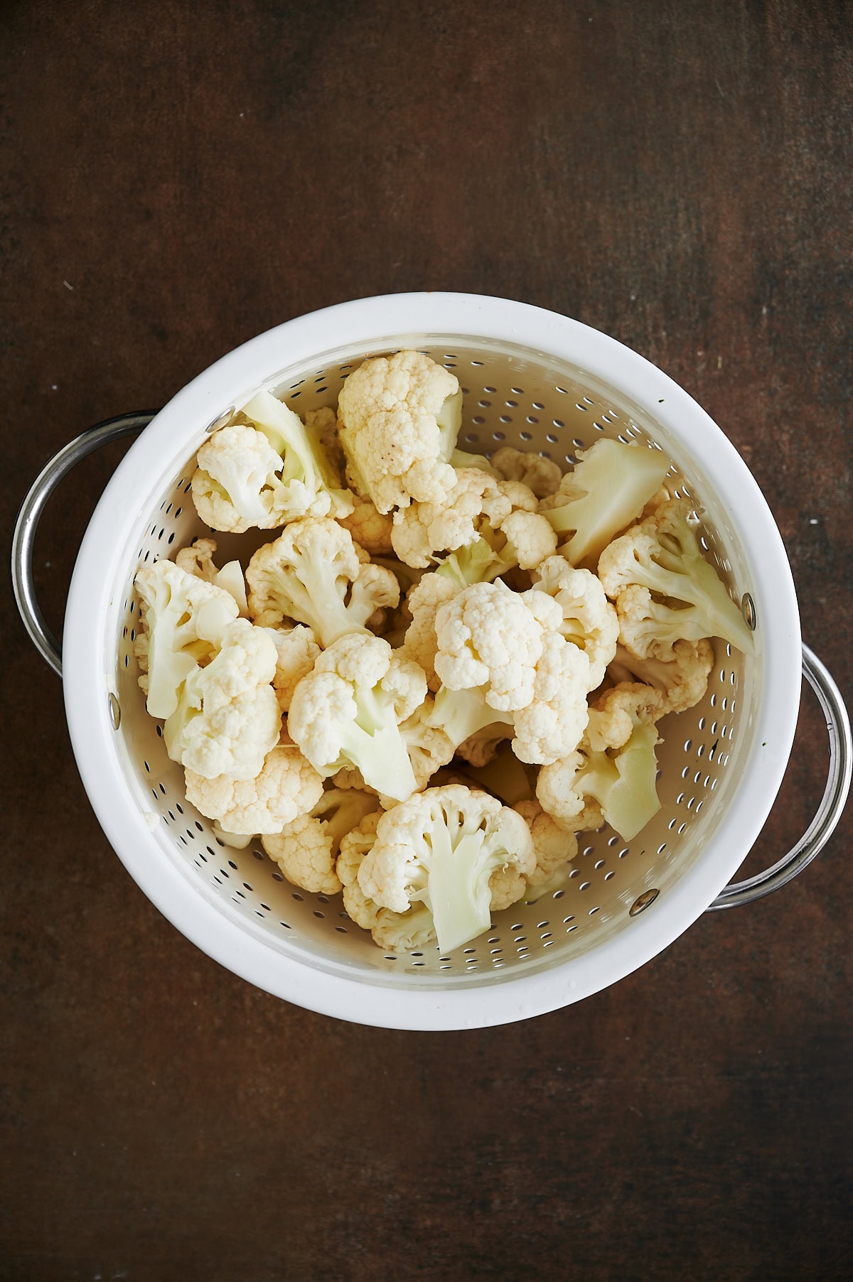 A white colander filled with raw cauliflower florets sits on a dark brown surface.
