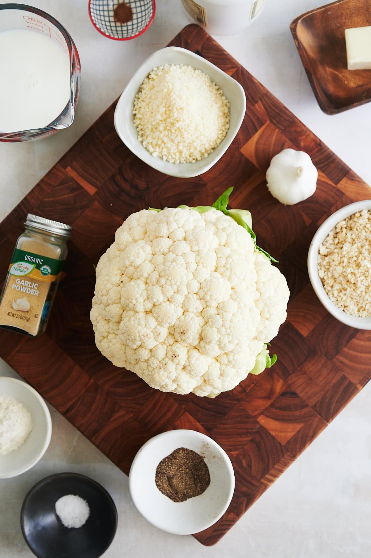 A head of cauliflower sits on a wooden cutting board surrounded by small bowls of milk, grated cheese, breadcrumbs, salt, pepper, garlic powder, and a whole garlic bulb.