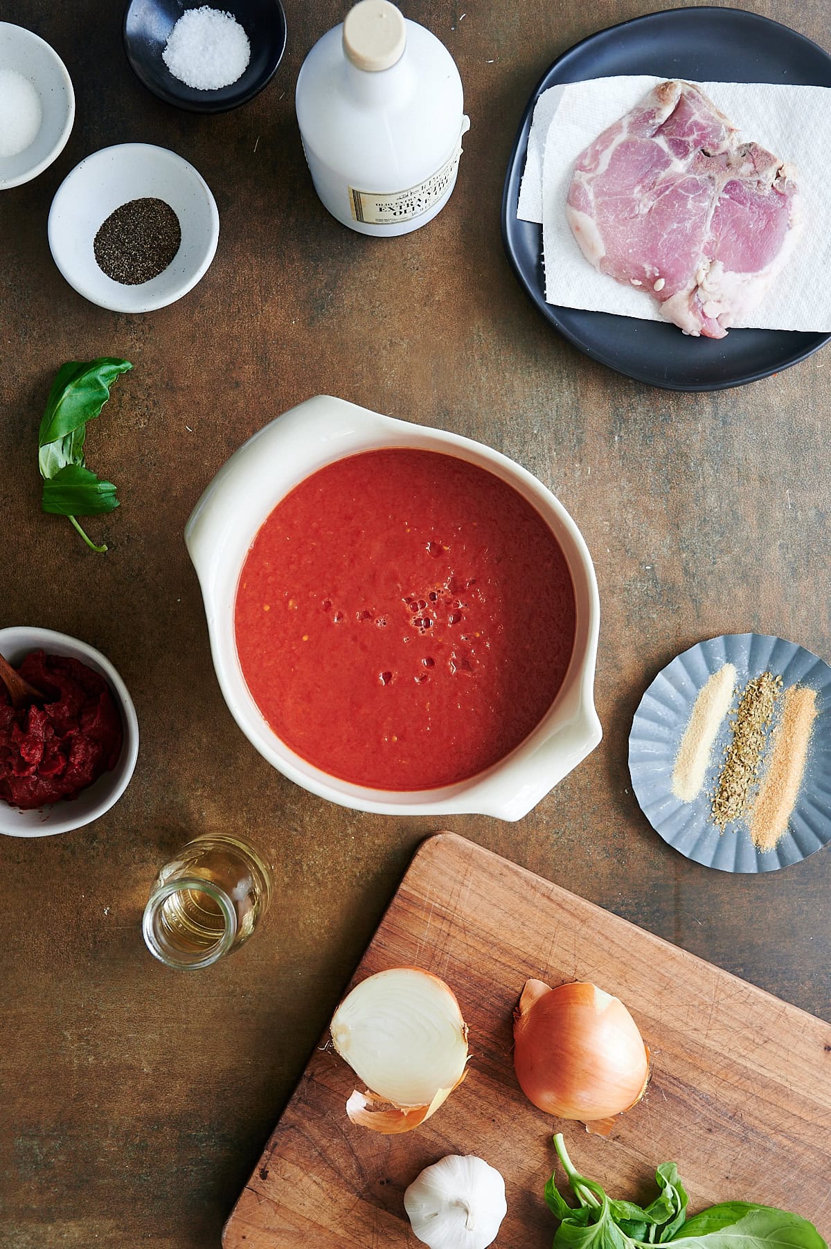 A bowl of whole tomatoes surrounded by raw pork, onions, garlic, basil, spices, salt, pepper, tomato paste, oil, and vinegar on a wooden surface.