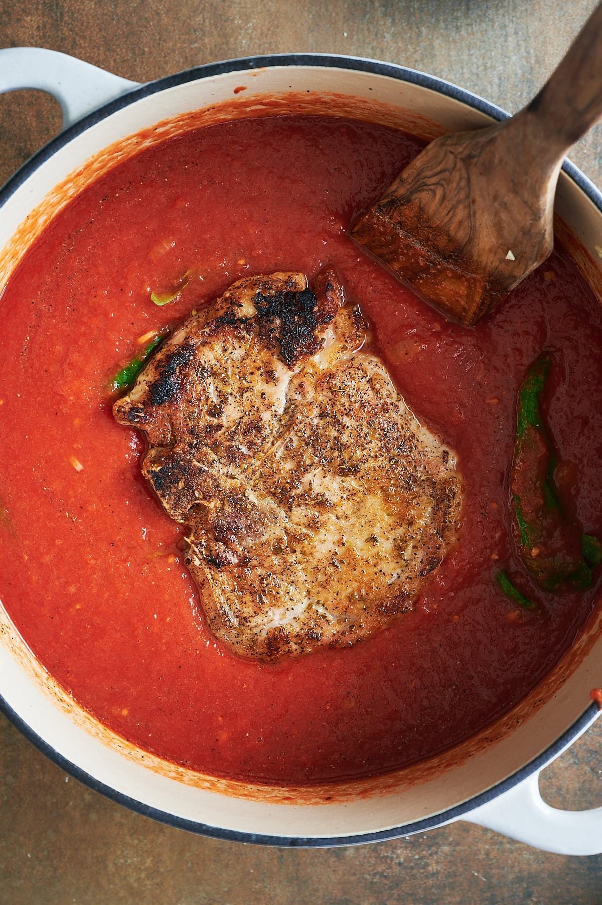 A seasoned pork chop cooking in a pot of tomato sauce, with a wooden spatula resting on the side.