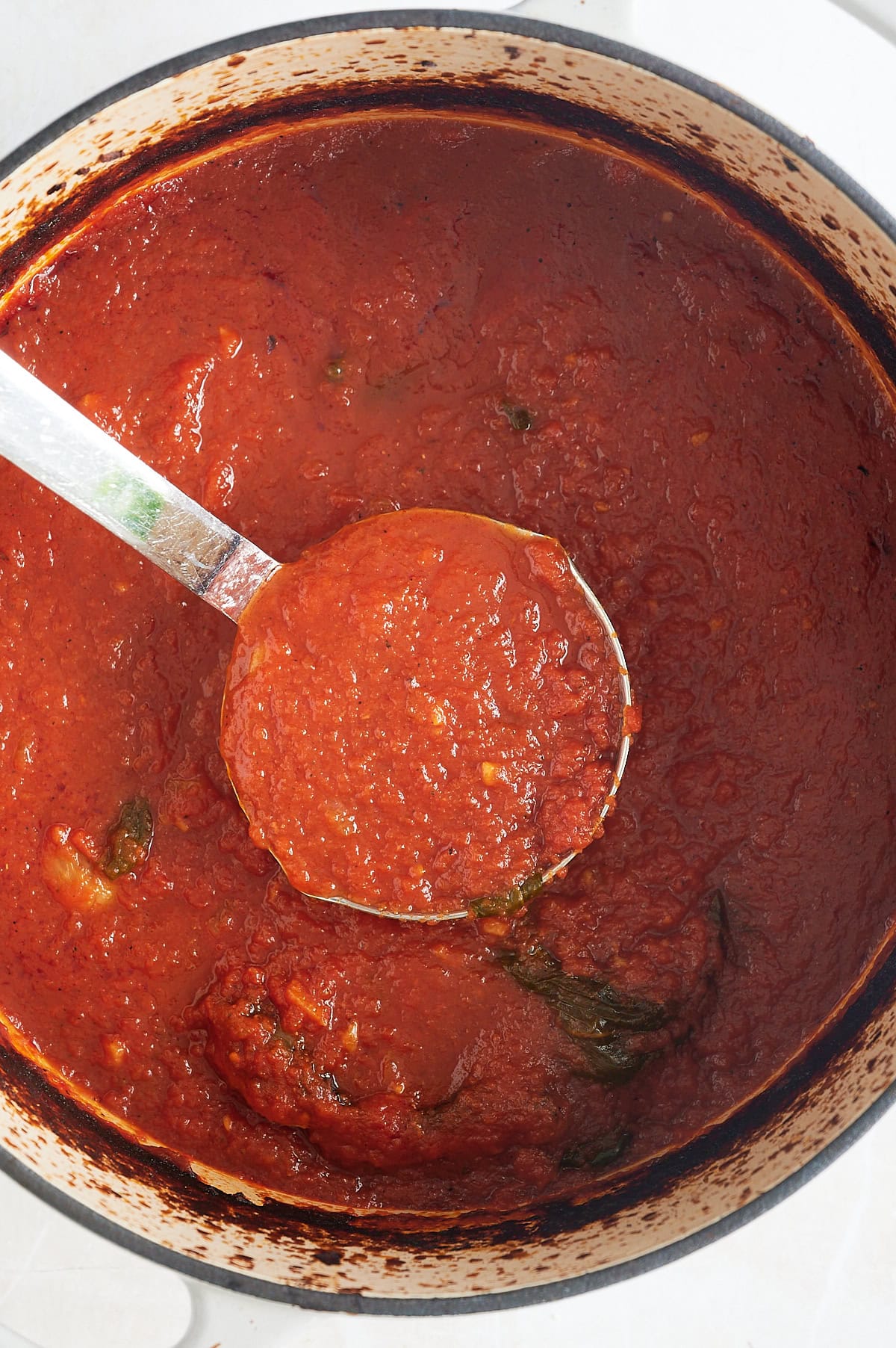 A ladle scoops tomato gravy from a large pot; the sauce is thick and red with visible herbs and small chunks of ingredients.