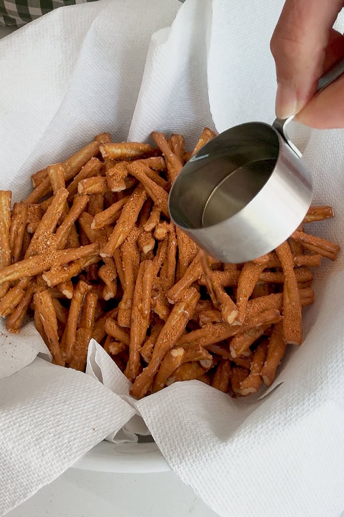 A hand pours oil from a metal measuring cup onto broken pretzel sticks in a paper towel-lined bowl.