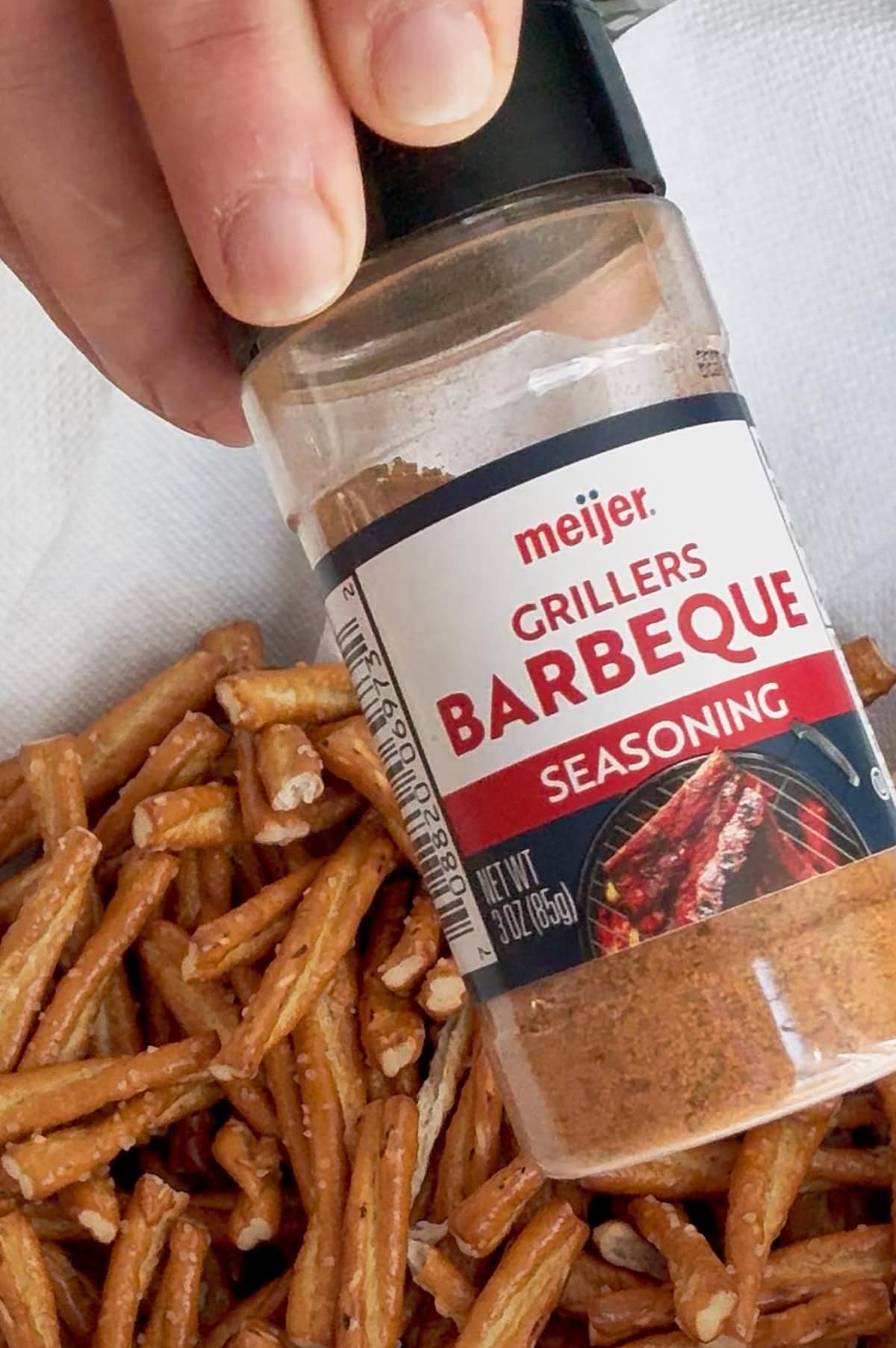 A hand holds a bottle of Meijer Grillers Barbeque Seasoning above a pile of pretzel sticks on a white surface.