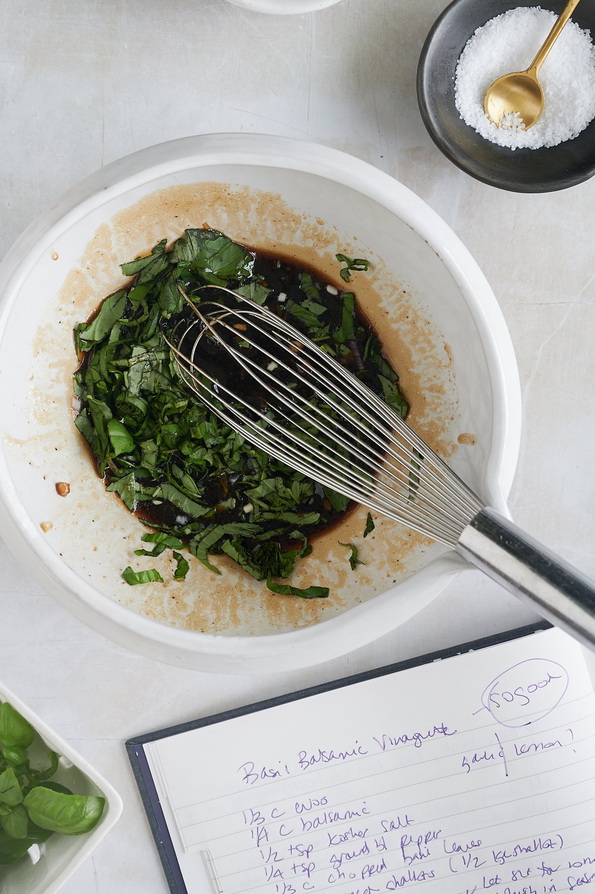 A bowl with chopped basil and balsamic vinegar is being whisked; a recipe notebook, salt bowl, and basil leaves are nearby on a white surface.