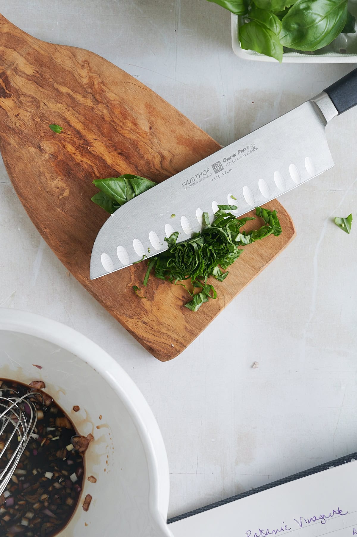 A chef’s knife with chopped basil sits on a wooden cutting board; nearby are a bowl with balsamic dressing and a bunch of fresh basil leaves.