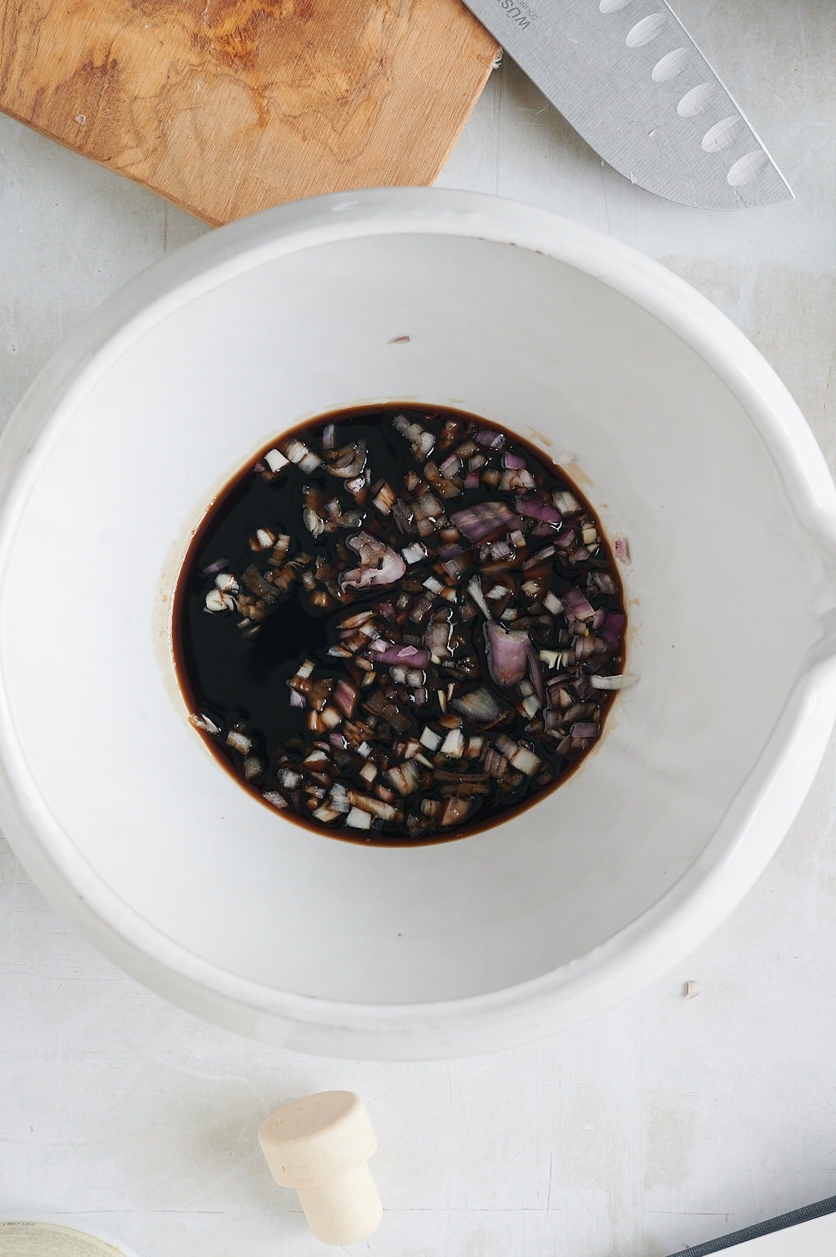 Chopped shallots and balsamic vinegar in a white mixing bowl on a light countertop with part of a knife and wooden board visible.