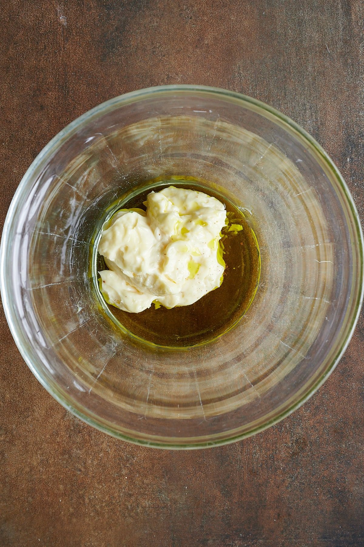 A glass bowl containing a mixture of mayonnaise and olive oil on a brown surface.