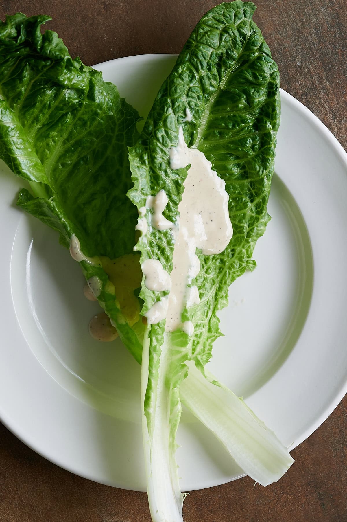 A white plate with two romaine lettuce leaves and a drizzle of creamy Caesar dressing on top.