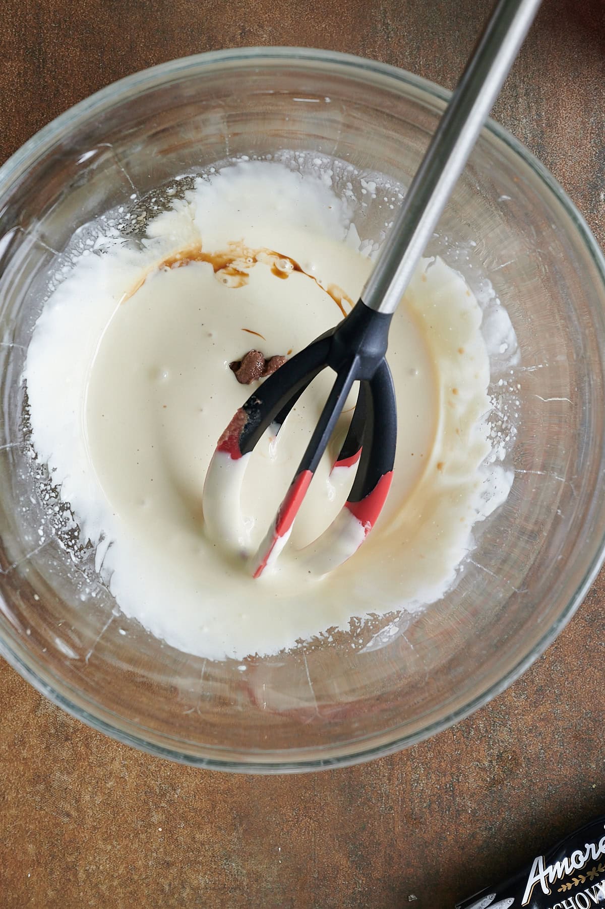 A glass bowl stirring anchovy paste into Caesar dressing base with a red and black whisk, on a brown surface.