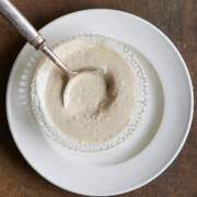 A metal spoon rests in a small glass bowl filled with creamy white sauce, placed on a white plate against a brown surface.