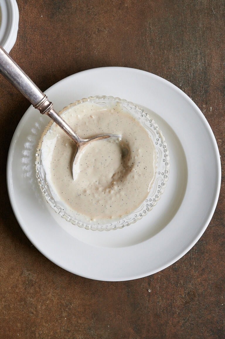 A metal spoon rests in a small glass bowl filled with creamy white sauce, placed on a white plate against a brown surface.