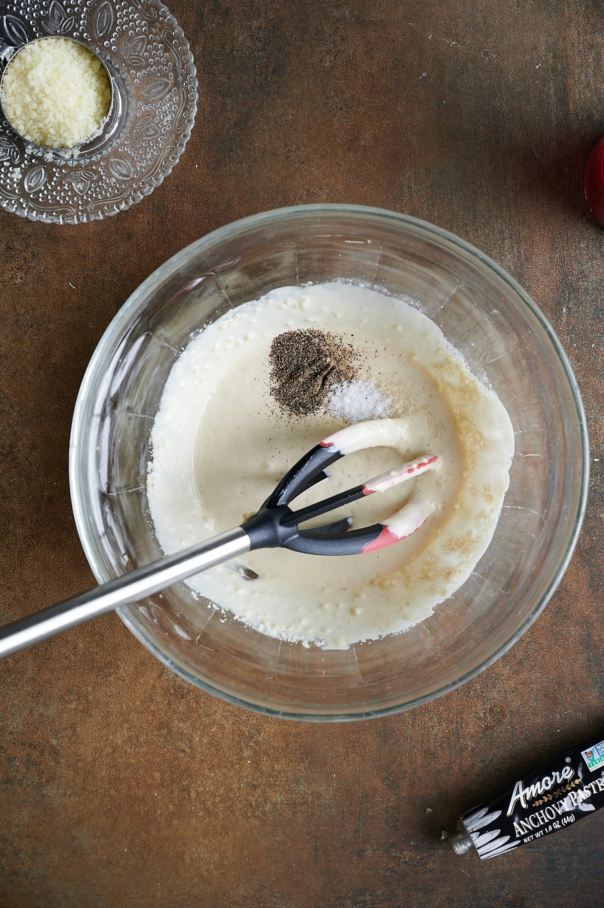 A glass bowl containing a creamy Caesar dressing base with ground pepper and salt on top, being mixed with a whisk; cheese and anchovy paste are nearby.