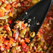 A close-up of a bowl containing a mixture of chopped green olives, red bell peppers, onions, and oil with a black spoon resting in the giardiniera.