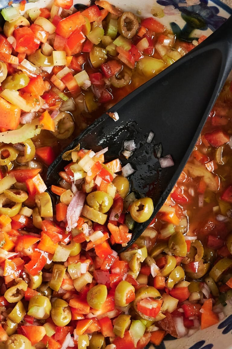 A close-up of a bowl containing a mixture of chopped green olives, red bell peppers, onions, and oil with a black spoon resting in the giardiniera.