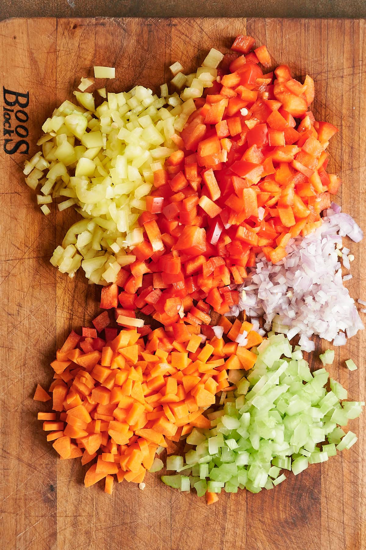 Diced yellow bell pepper, red bell pepper, shallot, celery, and carrot arranged on a wooden cutting board.