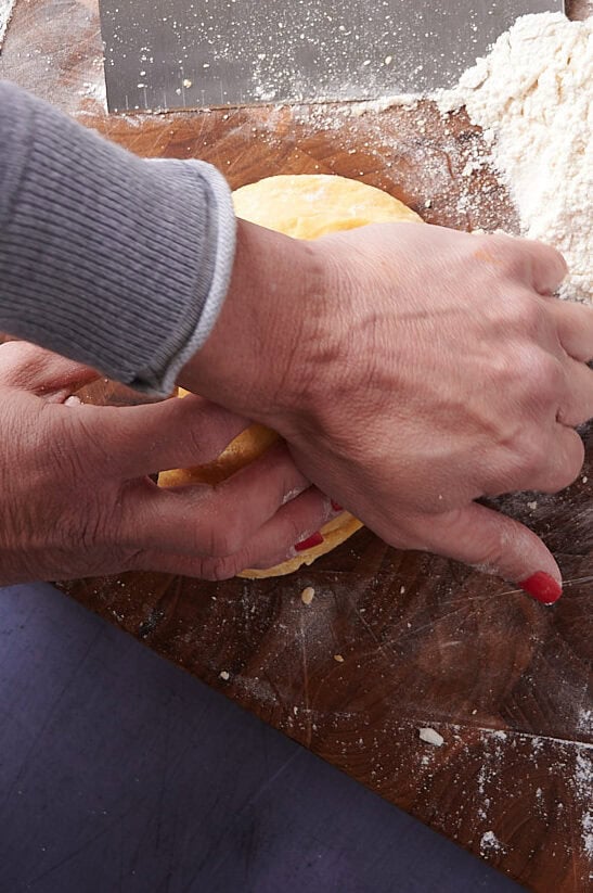 A person presses homemade ravioli dough on a floured wooden surface next to a dough scraper and a pile of flour.