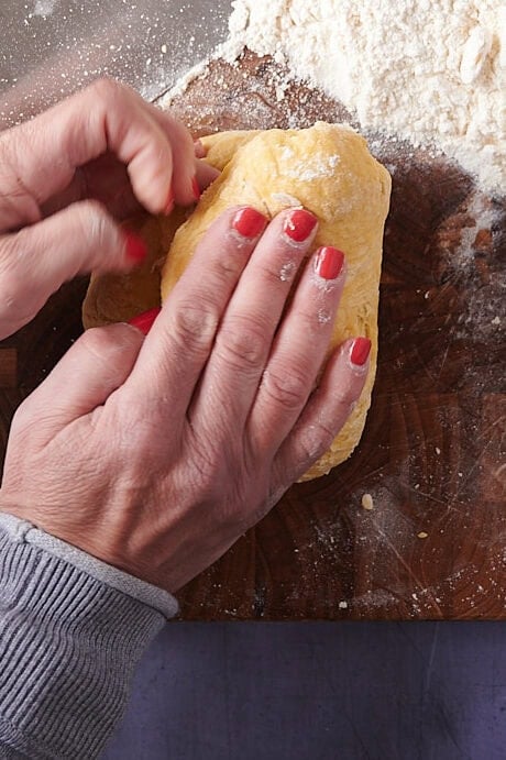 Top-down view of hands kneading homemade ravioli dough on a floured wooden cutting board, with a bench scraper and a pile of flour nearby.