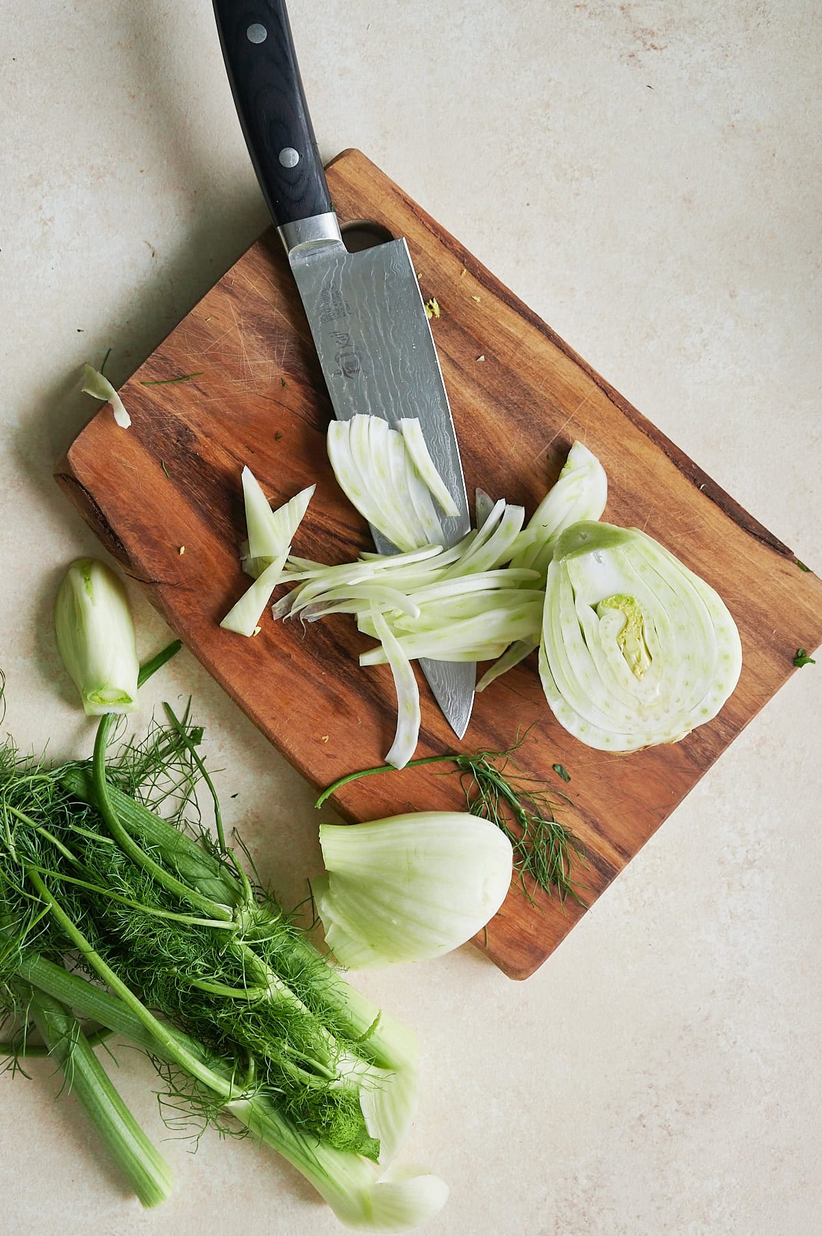 A fennel bulb is sliced on a wooden cutting board with a kitchen knife, surrounded by fennel fronds and pieces on a light countertop.