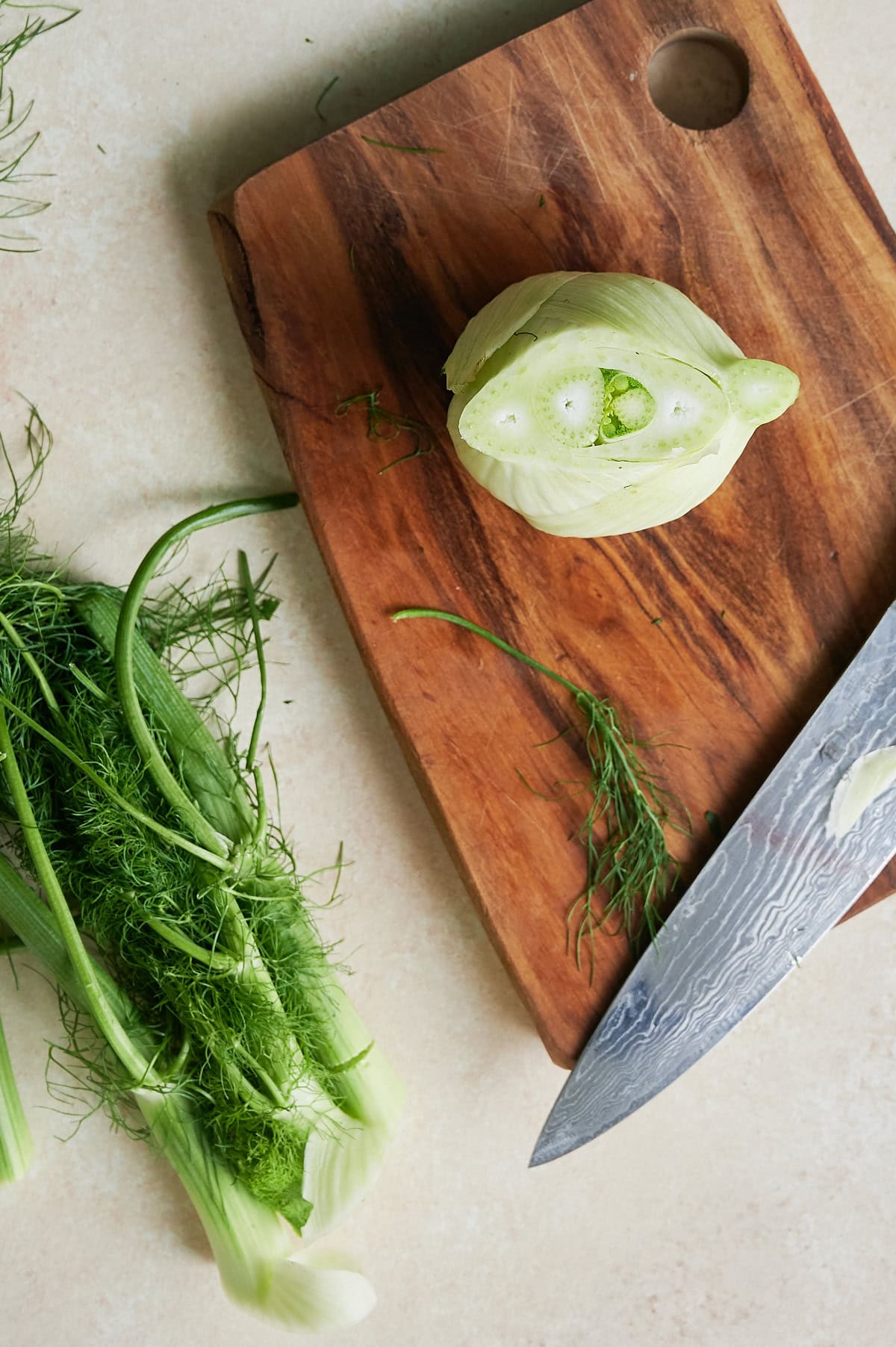 A fennel bulb on a wooden cutting board next to a knife and fennel fronds on a light countertop.