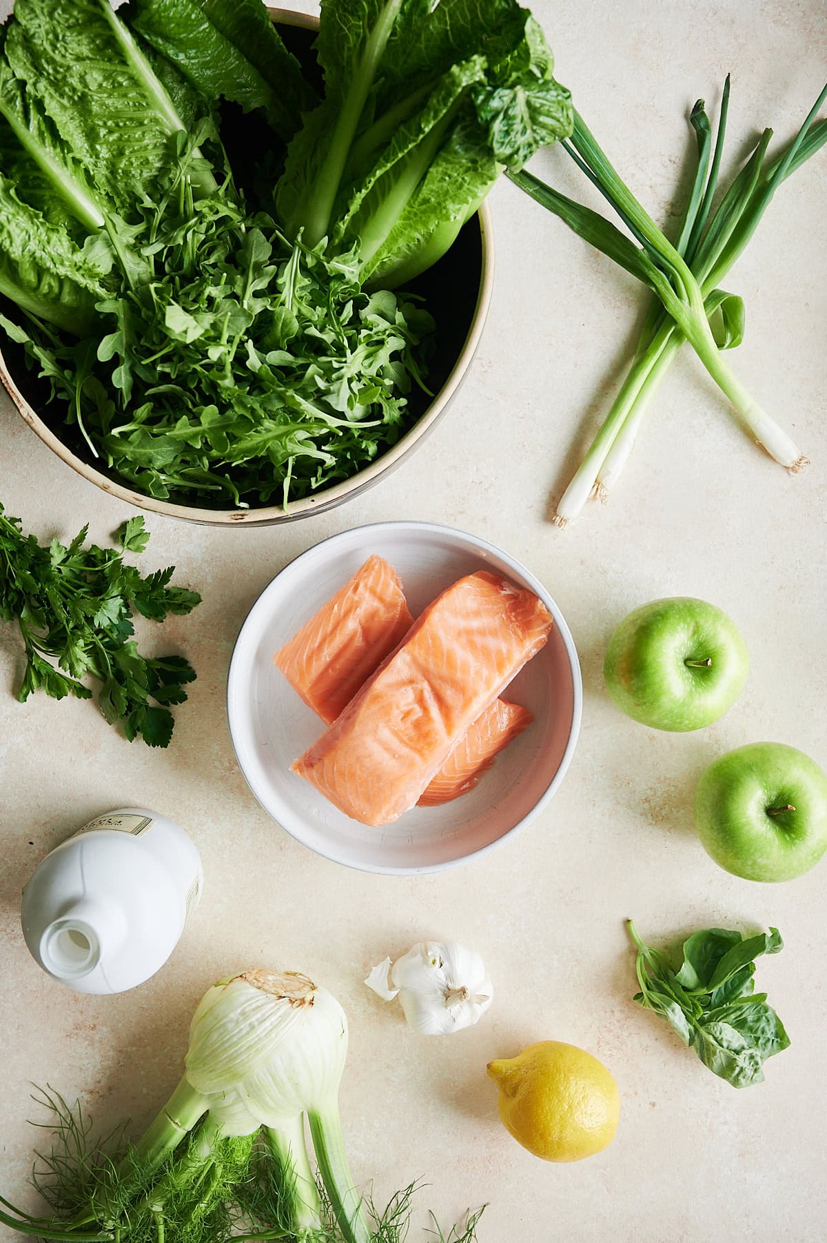 Fresh ingredients including salmon fillets, green apples, leafy greens, green onions, fennel, lemon, garlic, parsley, basil, and a bottle of oil arranged on a light surface.