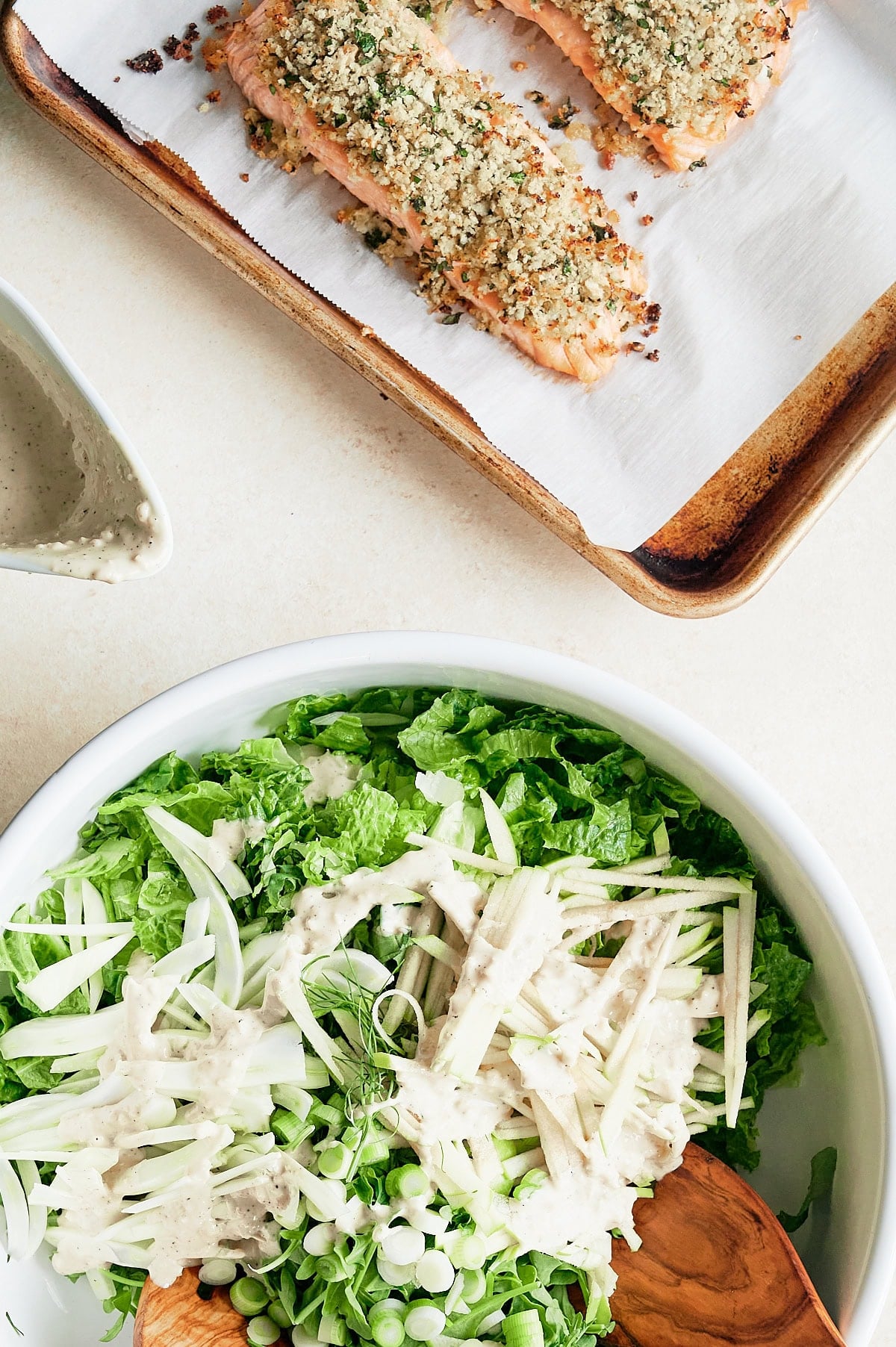 A baking tray with herb-crusted salmon fillets and a large bowl of green salad topped with creamy dressing and sliced vegetables.