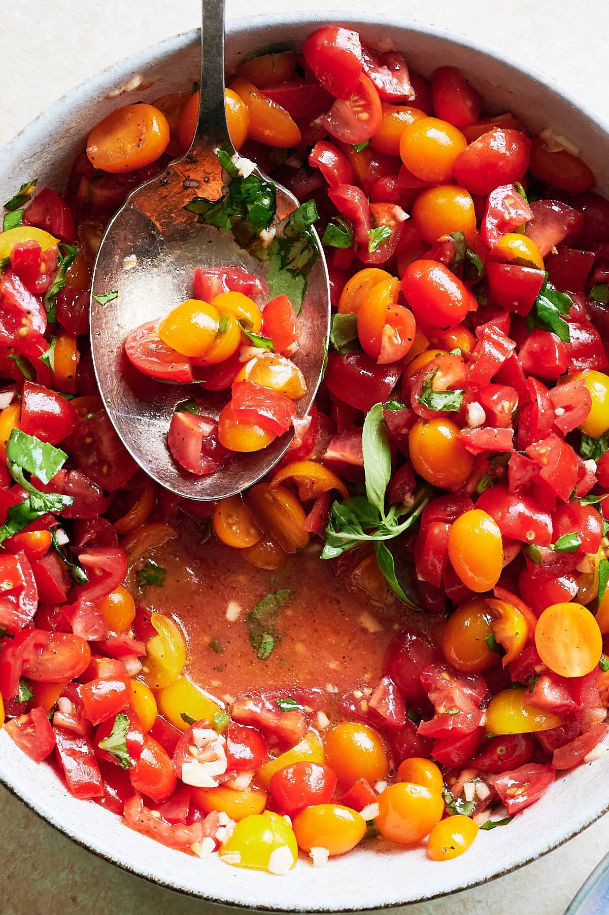 A bowl of chopped red and yellow cherry tomatoes mixed with fresh herbs and garlic, with a large spoon resting inside.