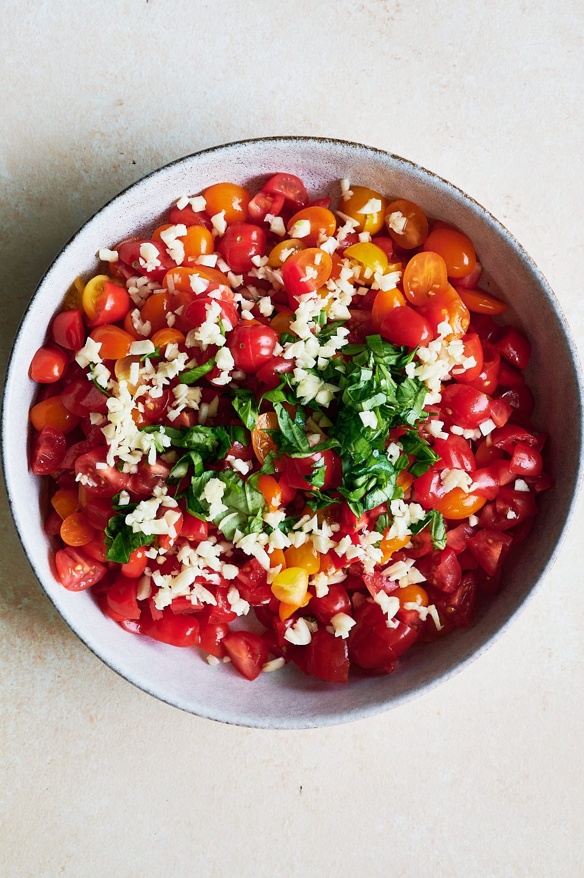 A bowl filled with bruschetta including chopped red and yellow cherry tomatoes, minced garlic, and fresh basil leaves on a light surface.