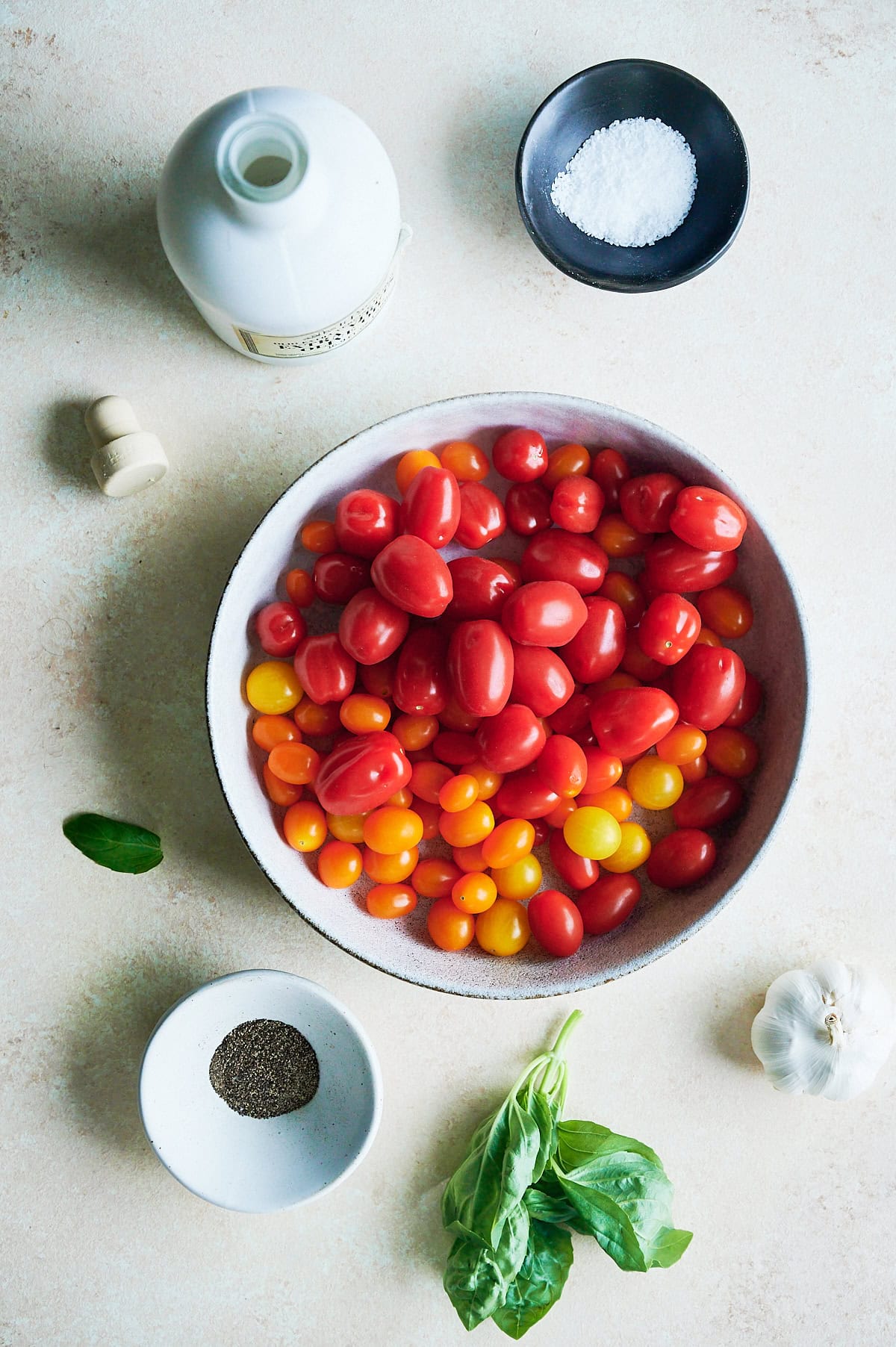 A bowl of red and yellow grape tomatoes surrounded by salt, pepper, olive oil, a garlic bulb, and fresh basil leaves on a light surface for bruschetta.