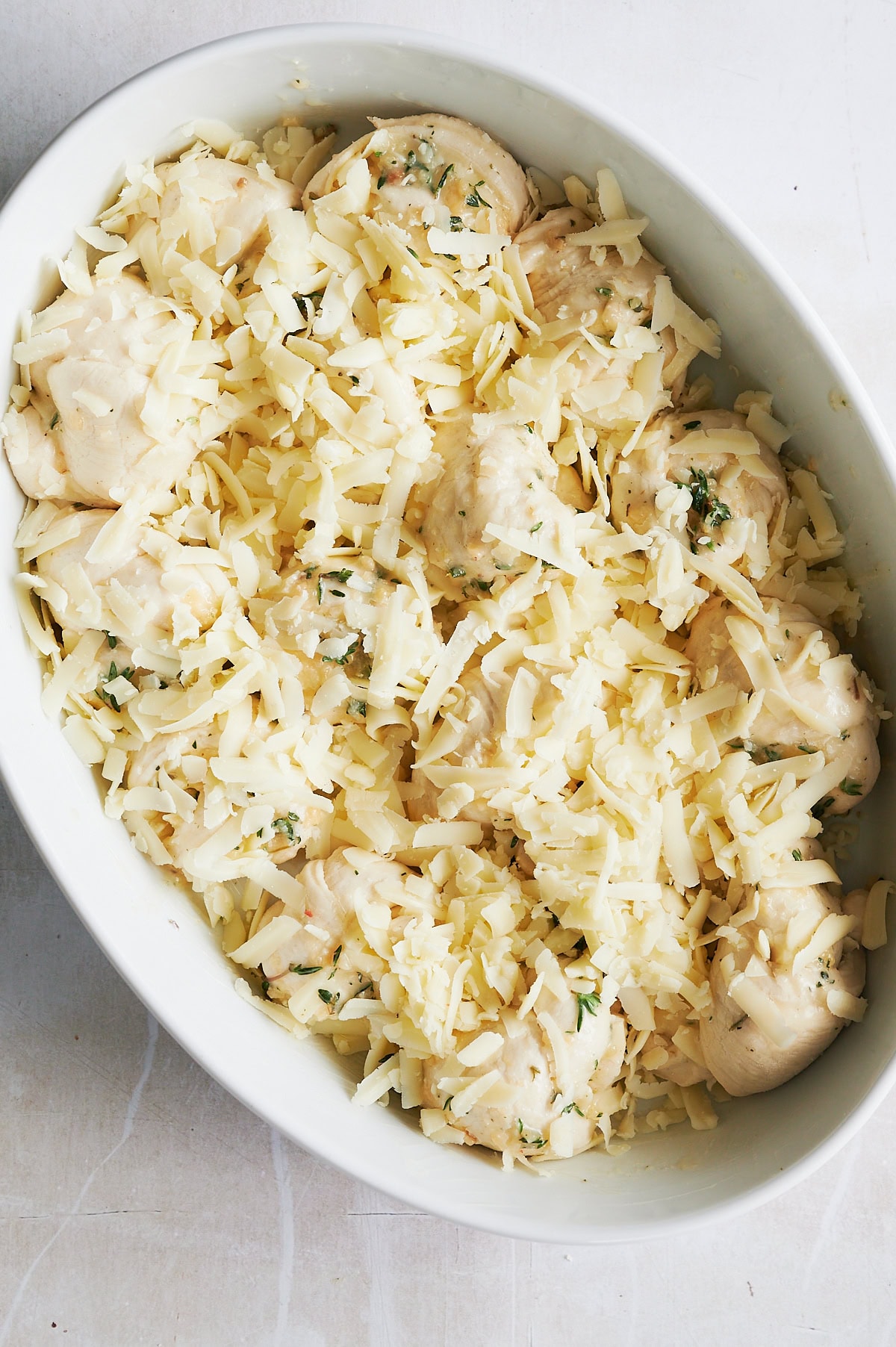 Oval white dish filled with uncooked stuffed mushrooms topped with shredded cheese and herbs, placed on a light-colored surface.
