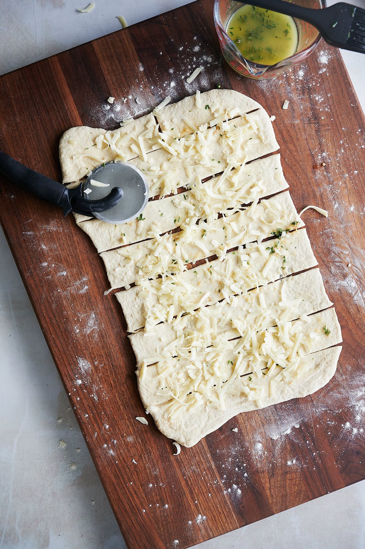 Rectangular pizza dough topped with shredded cheese and herbs is sliced into strips on a wooden board with a pizza cutter; a bowl of egg wash is nearby.