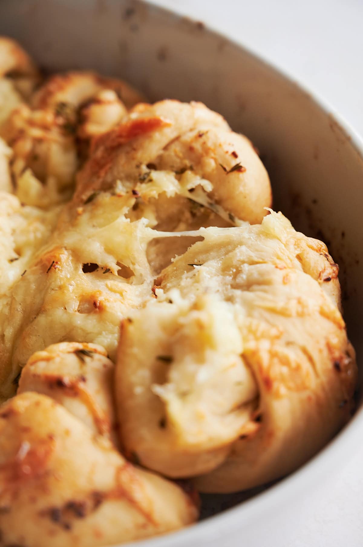 Close-up of freshly baked, golden brown pull-apart bread with melted cheese and herbs in a white baking dish.